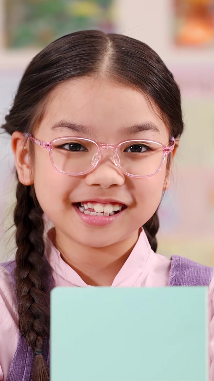 Smiling young girl in pink glasses reads a book inside a bright, colorful classroom