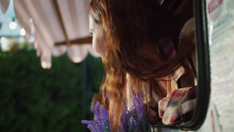 Happy-brunette-girl-in-a-plaid-shirt-sticks-her-head-out-to-the-city-and-looks-around-from-a-trailer-during-her-picnic-at-a-camp-outside-the-city-in-summer.-Happy-brunette-girl-looking-out-of-trailer-in-summer