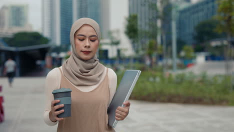 a woman in a hijab walking in the city with a coffee and laptop