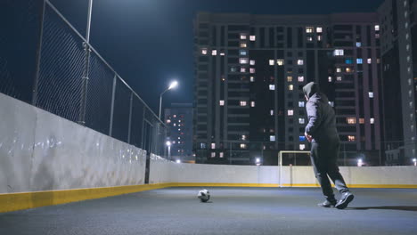 side view of man in hoodie playing soccer under bright stadium lighting at night, ball near goalpost with reflection on wall, surrounded by urban cityscape and illuminated office buildings