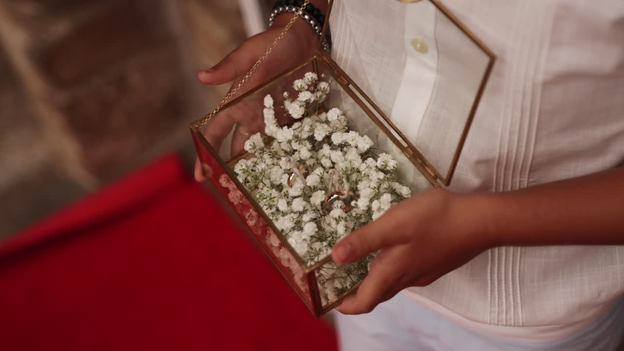 Child holding a glass box with gold wedding rings and white flowers during ceremony