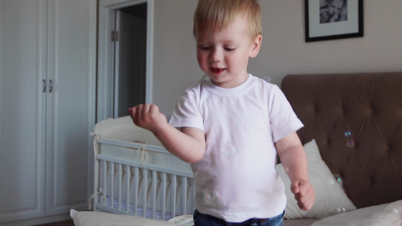 A boy in a white T-shirt and blue jeans catches soap bubbles standing on the bed in the parent bedroom