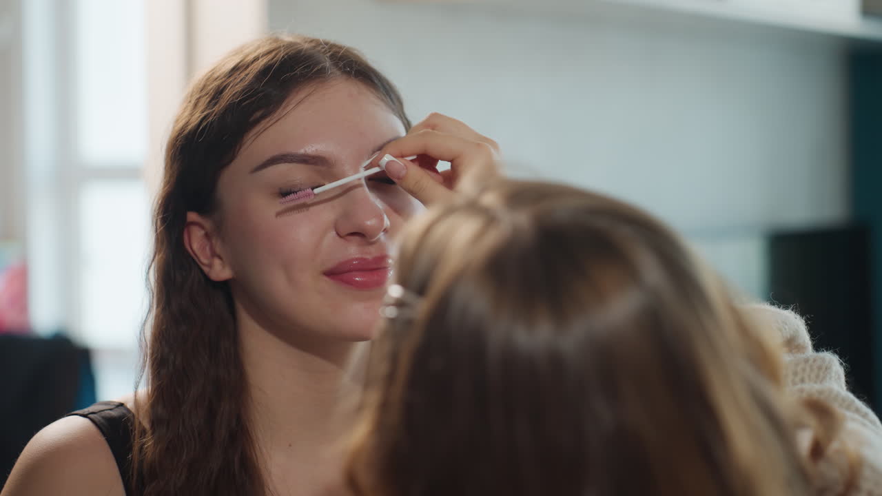 Model Undergoing Lash Grooming In Studio, Makeup Artist Carefully Applying Mascara On Seated Model, Caucasian Woman Being Beautified With Mascara And Lash Grooming In Softly Lit Studio Setting
