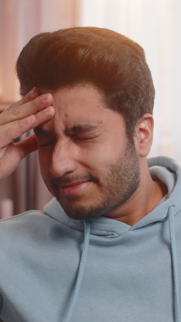 Indian young man on sofa rubbing temples to cure headache suffering from stress and migraine at home