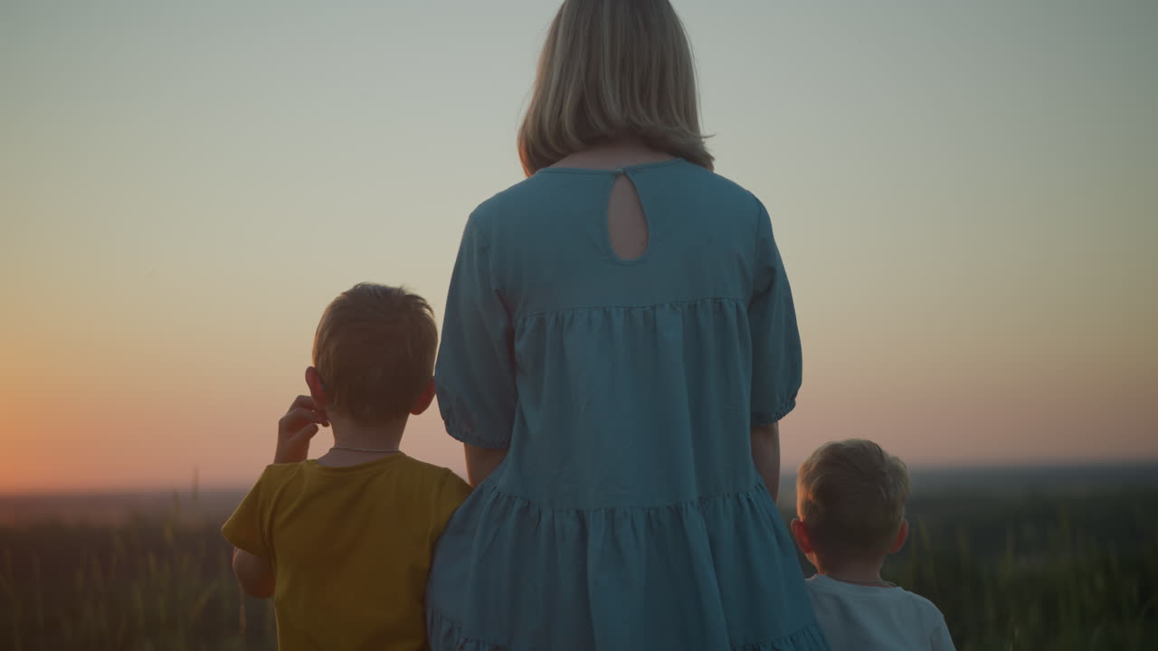 A close-up back shot of a mother in a blue gown with her two young sons, one wearing yellow and the other in white. They stand together, gazing at a peaceful sunset over a calm lake