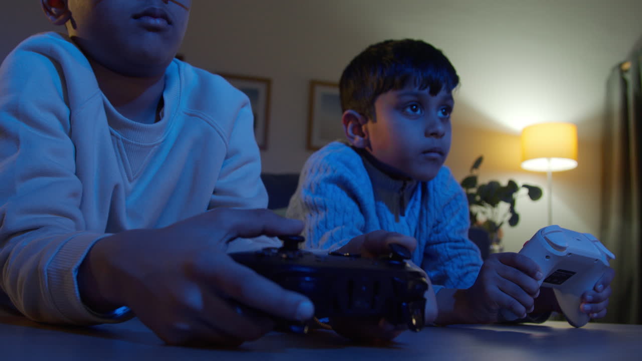 Two Young Boys At Home Playing With Computer Games Console On TV Holding Controllers Late At Night 6