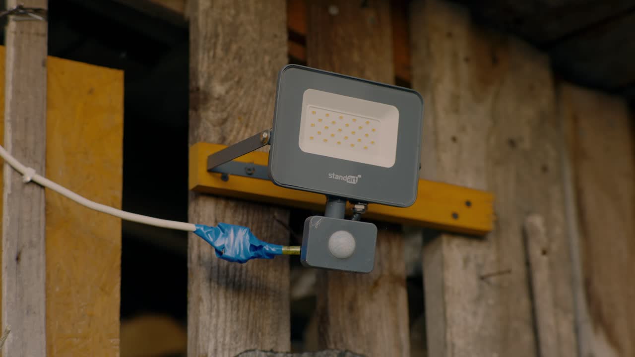 LED floodlight fixture mounted on a rustic wooden wall with a taped wiring connection. The industrial look and makeshift repair convey a practical, outdoor lighting setup.
