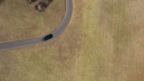 an aerial, top down drone view over a single car driving through an empty park on a sunny day