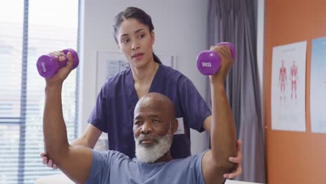 una fisioterapeuta femenina diversa y un paciente masculino anciano sosteniendo pesas en una sesión de fisioterapia.