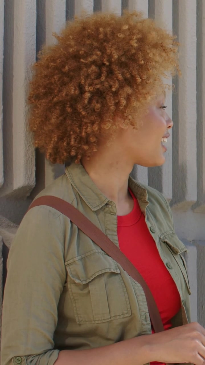 Video verticale: Donna che gira la testa mentre il sole le illumina il viso, arricciando le labbra vicino a una parete a coste, giacca color oliva.