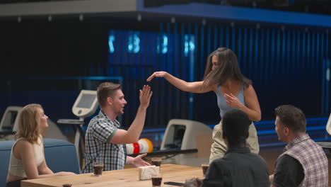 una mujer morena alegre corre a los amigos dando cinco hombres y mujeres sentados en una mesa en un club de bolos. celebrar la victoria y la racha. para cortar y felicitar por la victoria. éxito del equipo.