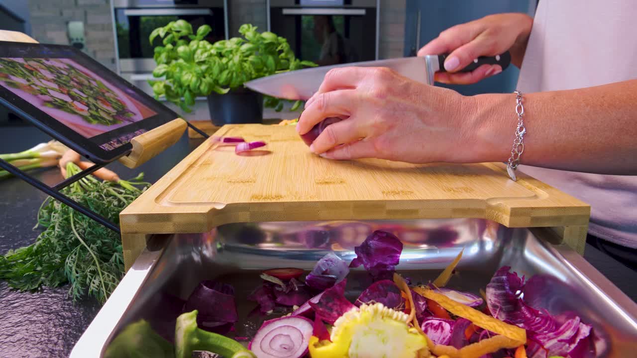 primer plano panorámico de una mujer cortando verduras en una tabla de cortar con una receta en una tableta en una cocina moderna y hierbas y verduras frescas