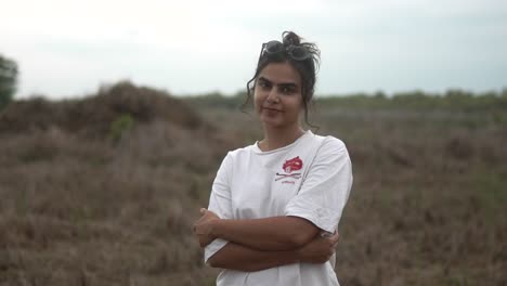 Mujer-Joven-Parada-Con-Confianza-En-Un-Campo-Con-Los-Brazos-Cruzados,-Sonriendo-Suavemente-A-La-Cámara.