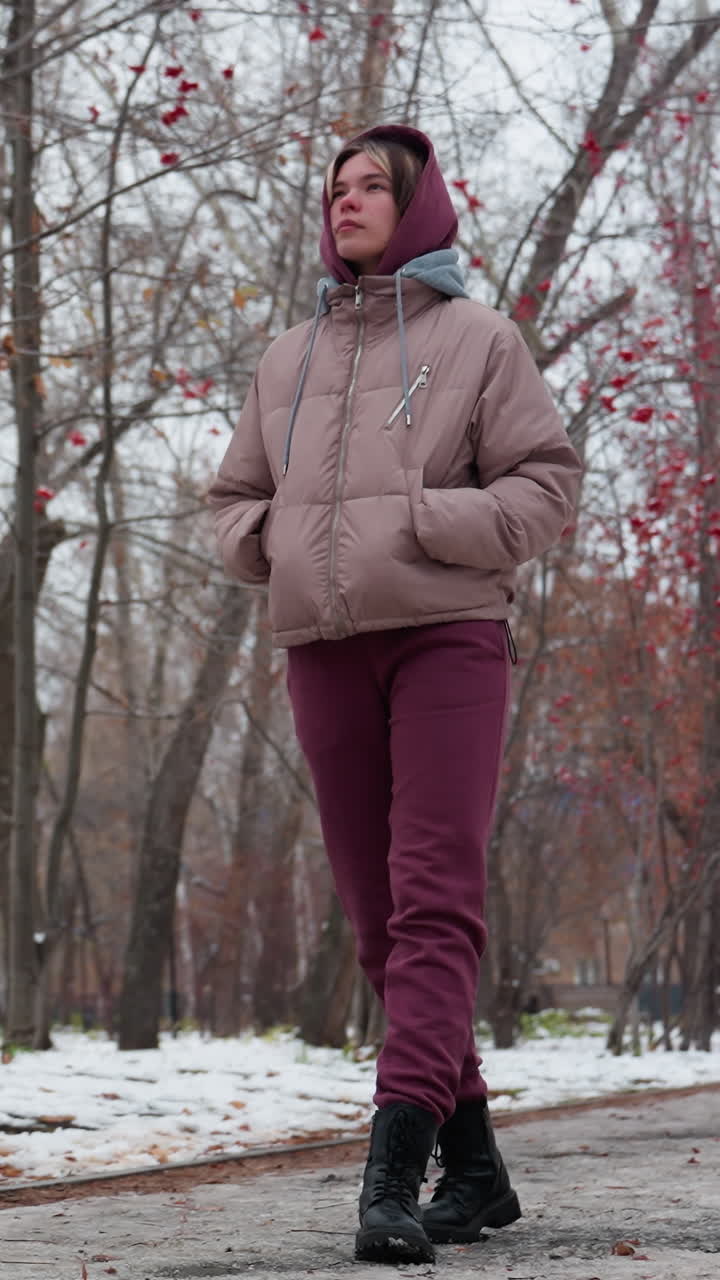 mujer en abrigos cálidos de invierno caminando con las manos en los bolsillos en una calle vacía nevada alineada con árboles desnudos adornados con bayas rojas, huellas visibles en el camino congelado