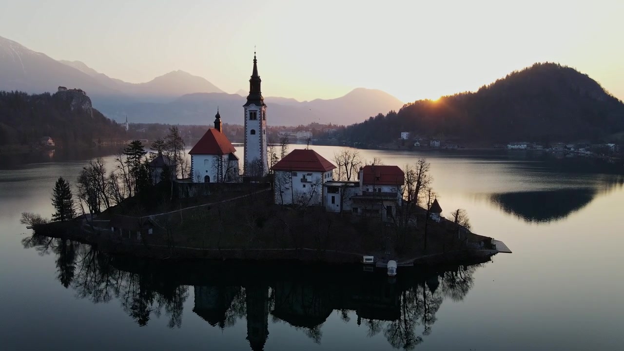 Drohne steigt über der Bleder Insel auf und fängt die Spiegelung der Kirche sowie den Glanz des Sonnenaufgangs ein – 4K.