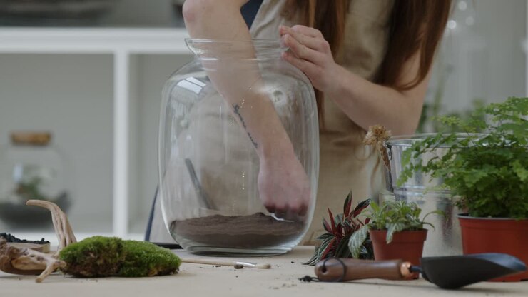 A young female florist prepares the first layer of soil in a glass jar for creating a tiny live forest ecosystem - close-up