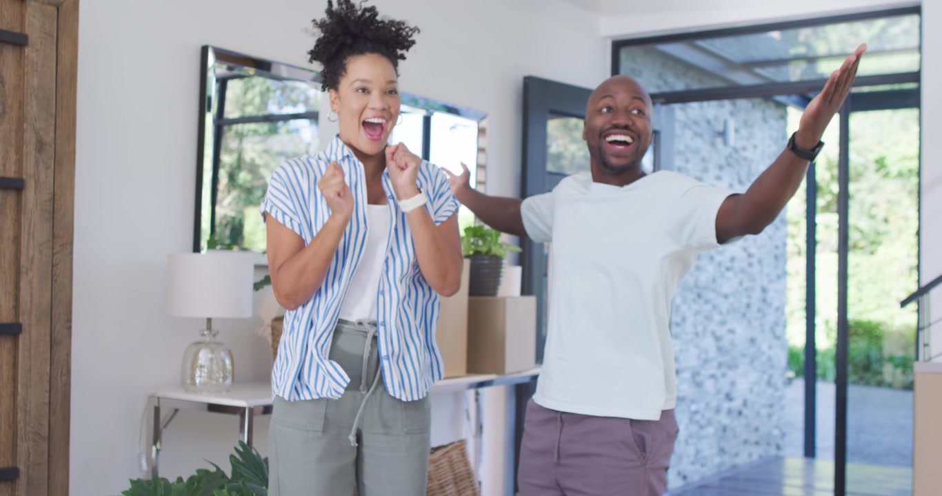 African American couple carrying boxes, woman holding key fob, man clapping, hugging in entryway