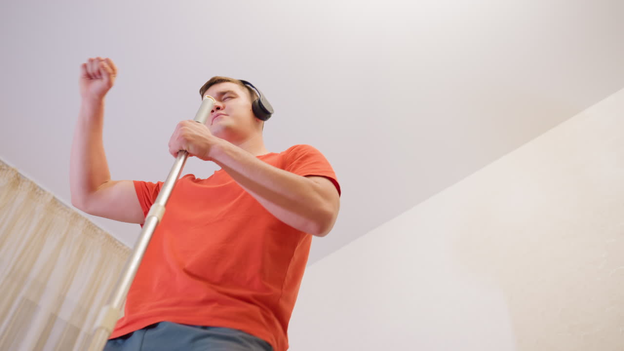 Giovane uomo con camicia arancione e cuffie, visto dal basso mentre balla e pulisce, con un'espressione che denota piacere nell'ascoltare musica durante i lavori domestici