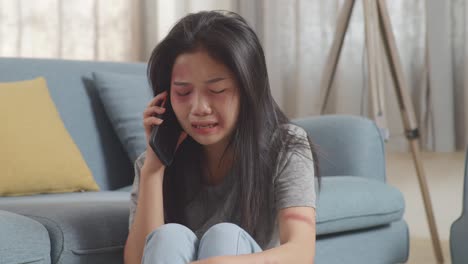 close up of young asian woman victim of violence with bruise on body crying and talking on smartphone asking for a help while sitting on the floor at home