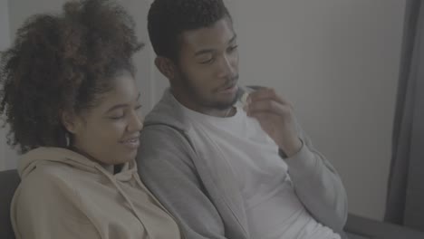 young african american couple having fun while watching a movie on tv. he gives his girlfriend a popcorn to eat