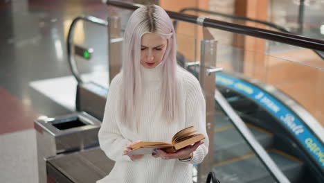 high angle view of shopper seated on mall bench flipping book pages with focused gaze wearing white sweater and jeans black tote bag beside modern glass railing escalator background polished floor