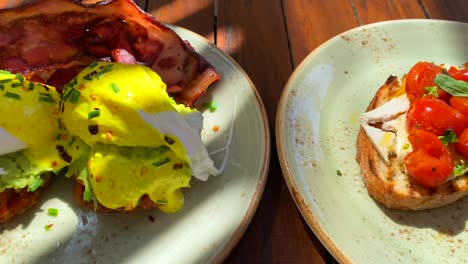 Leckeres-Eggs-Benedict-Frühstücksgericht-Mit-Knusprigem-Speck,-Sauce-Hollandaise-Und-Avocado-Auf-Süßkartoffelrösti,-Bruschetta-Toast-Mit-Kirschtomaten,-Pochierte-Eier-Zum-Brunch-In-Einem-Restaurant,-4k