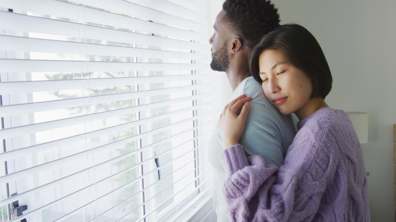 Happy diverse couple embracing and looking through window in bedroom