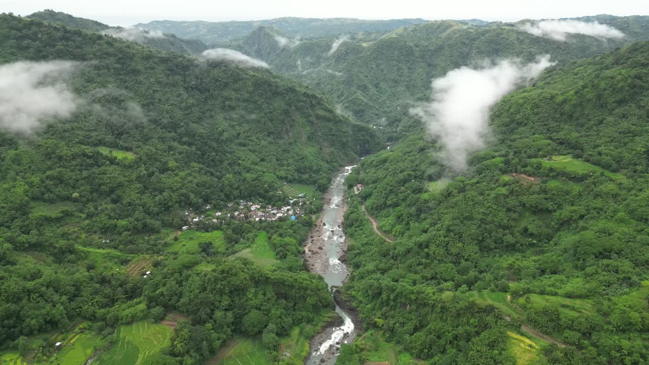 A wide aerial angle drifts to a final hold over forested mountains and a winding river. Terraced fields and a clustered village settle into the valley as mist threads through the rugged terrain