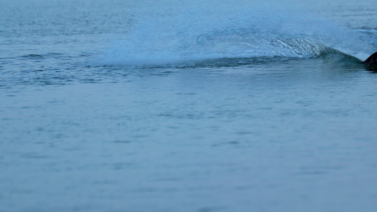 A person's foot, wearing a shoe, is skimming or stepping on the water's surface, creating a splash and a trail of water disturbance