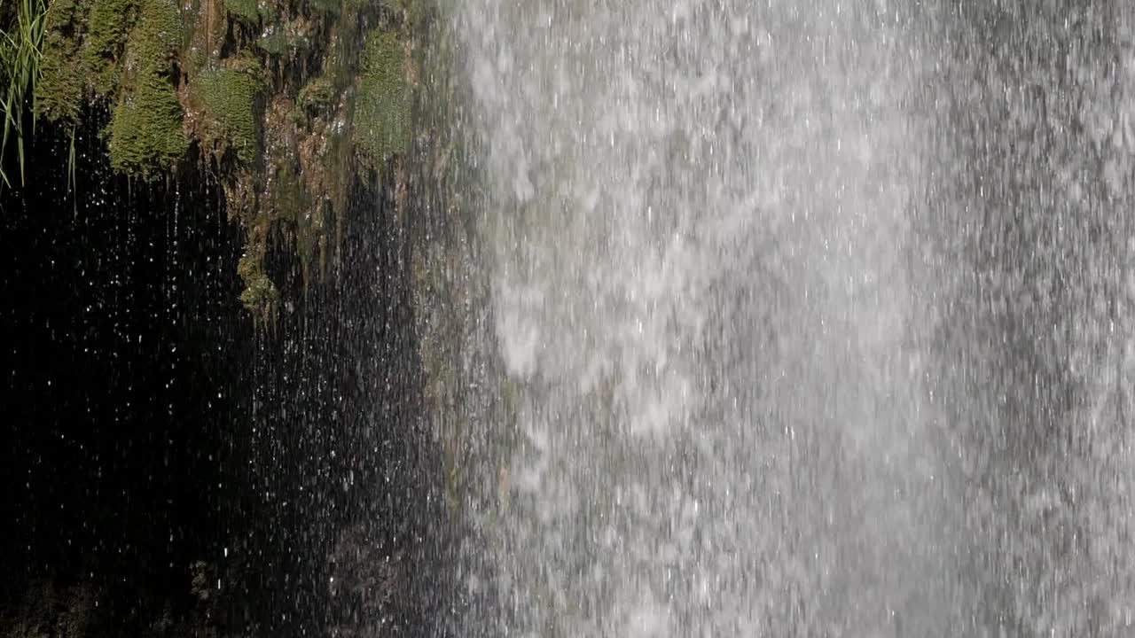 Closeup water curtain from waterfall cataract. Body of water fall strongly next to rock formation covered on moss. Dark cave gap