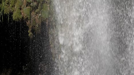 closeup water curtain from waterfall cataract. body of water fall strongly next to rock formation covered on moss. dark cave gap