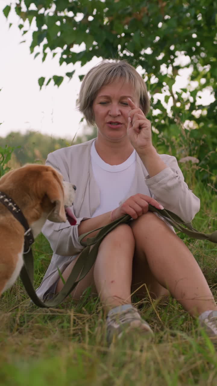 잔디에서 야외에 앉아있는 애완동물 애호가, 줄에 인 개들과 상호 작용하며, 왼쪽에 있는 개가 더 가까이 다가오는 동안 왼쪽의 개가 혀를 꺼내서 지켜보면서 한 마리를 장난스럽게 조롱합니다.