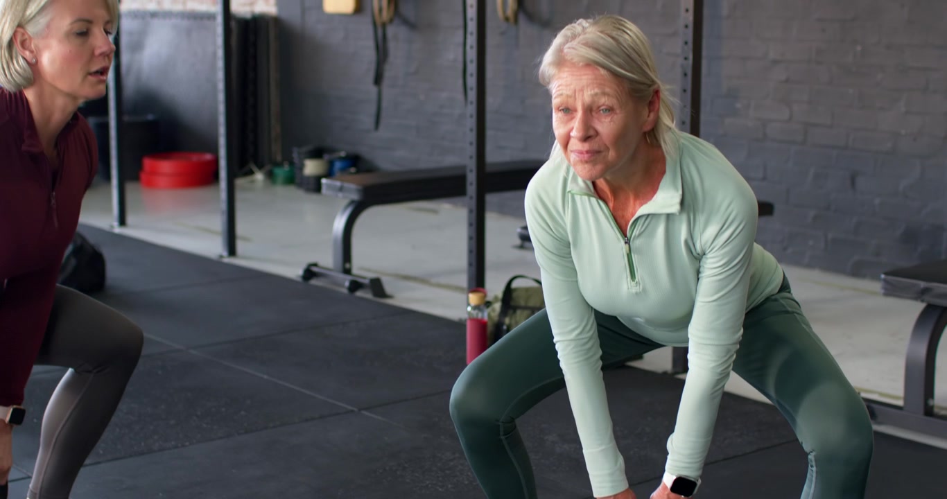 Istruttrice donna che istruisce una donna anziana con un top verde che fa oscillazioni con kettlebell in palestra per la forma.