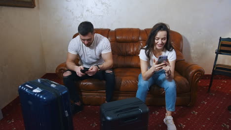 Happy young tourist couple indoors. Pretty woman and a man sitting on sofa in hotel near their suitcases and looking into phones.