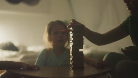 mom plays board games with cheerful toddler son and daughter sitting at small table in free time. family has fun playing and talking. family evening leisure