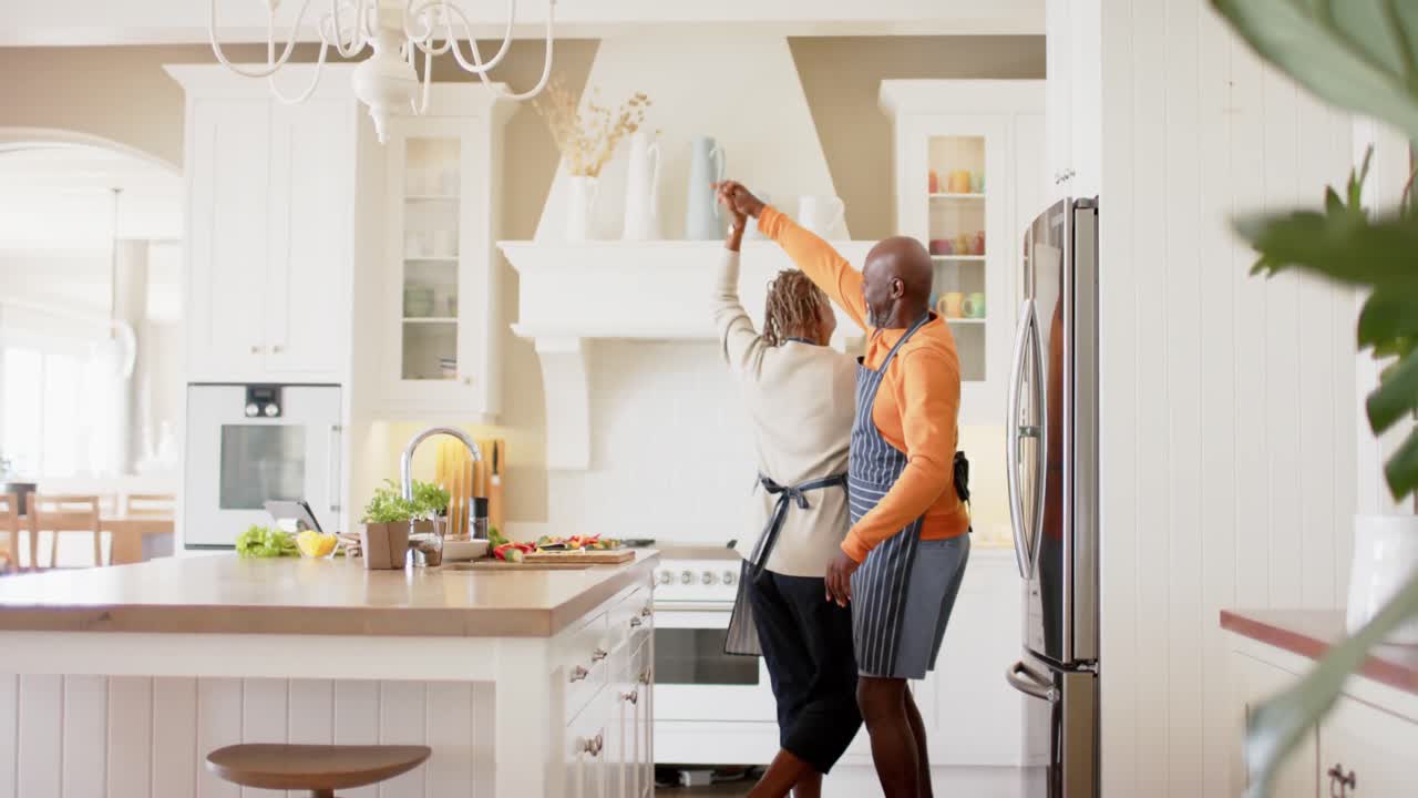 Happy african american senior couple in aprons dancing in kitchen, slow motion