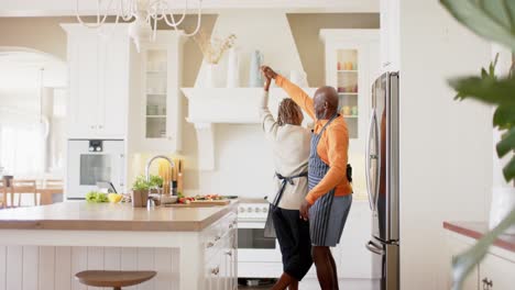 happy african american senior couple in aprons dancing in kitchen, slow motion