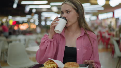 lady in pink dress picks up her drink, takes a sip with a satisfied expression, and places it back on the table, fries and burger are placed on a tray, with a blurred view of people in the background