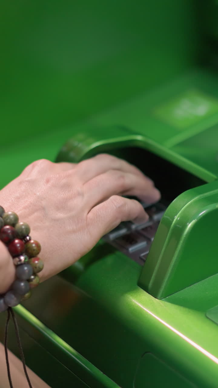 vista de una mano con pulseras de cuentas y una camisa negra escribiendo un alfiler en un cajero automático verde. tomada con una cámara de mano, capturando el momento de ingresar información bancaria