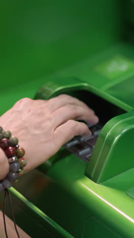 vista de una mano con pulseras de cuentas y una camisa negra escribiendo un alfiler en un cajero automático verde. tomada con una cámara de mano, capturando el momento de ingresar información bancaria