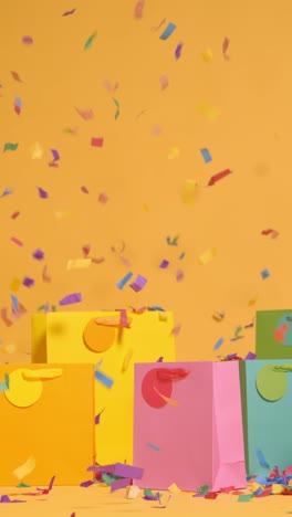 Vertical-Video-Studio-Shot-Of-Colourful-Birthday-Party-Gift-Bags-Against-Yellow-Background-With-Falling-Paper-Confetti