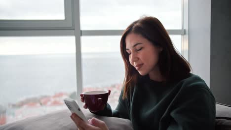 Happy-and-busy-brunette-girl-in-a-dark-green-sweater-typing-on-her-mobile-phone-and-holding-a-brown-mug-of-tea-in-a-modern-apartment-while-sitting-on-the-sofa