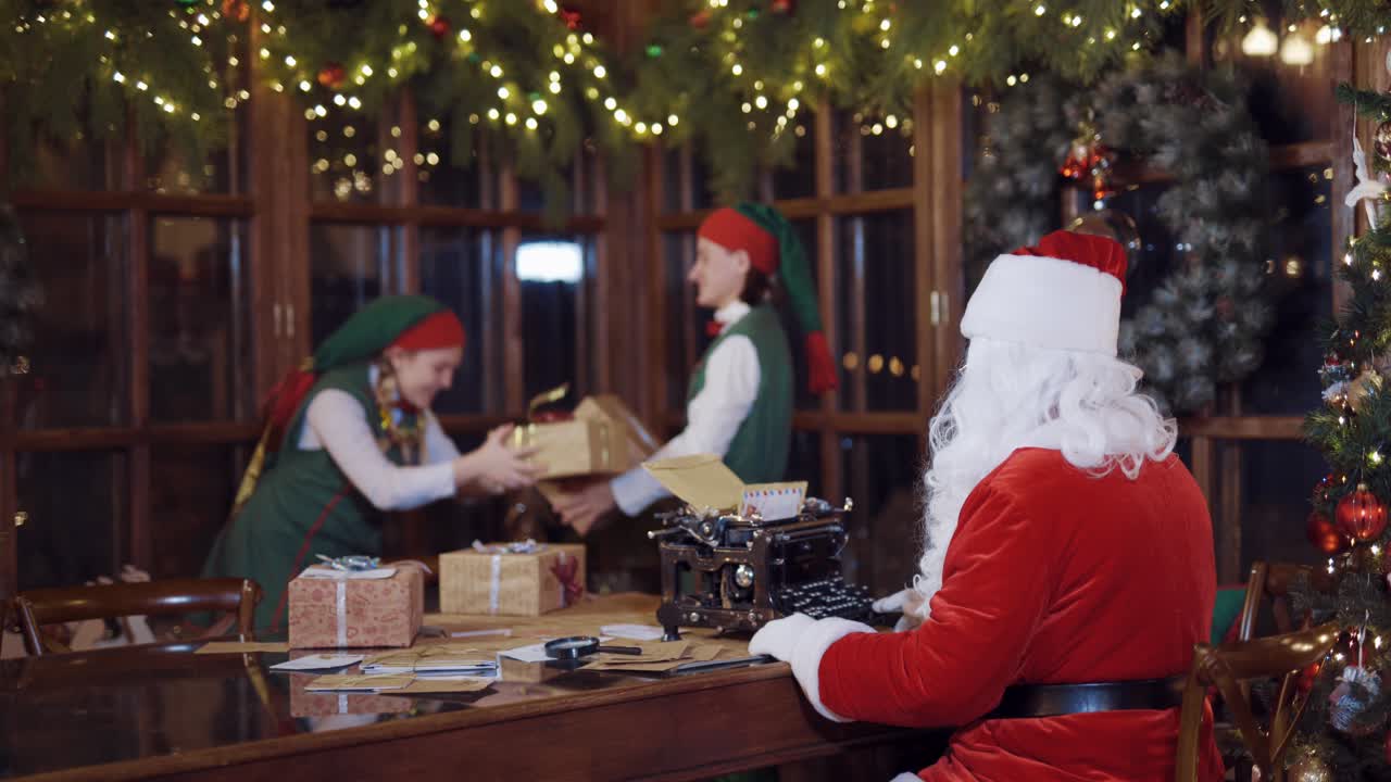 Joyful elves in green fairy suits play with boxes. Santa Claus is typing at the table while happy elves having fun with presents on Christmas decoration background.