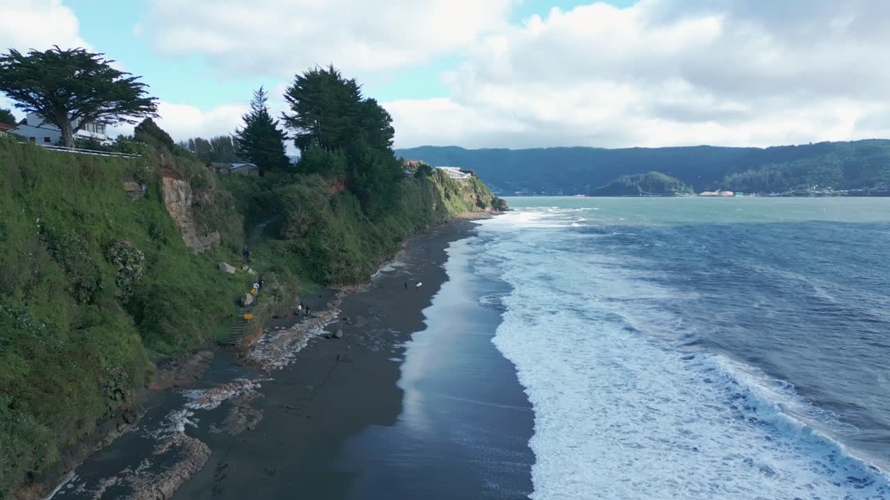 Drone-luftfoto fremover av Niebla svart sandstrand med hvitt skum, steinete klipper, skogkledde åser og panoramautsikt over Stillehavskysten