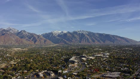 Toma-Aérea-De-Un-Amplio-Paisaje-De-Drones-De-Las-Impresionantes-Montañas-Rocosas-Cubiertas-De-Nieve-De-Utah-Con-El-Condado-De-Salt-Lake-Debajo-Lleno-De-Edificios-Y-árboles-Coloridos-En-Un-Cálido-Y-Soleado-Día-De-Otoño