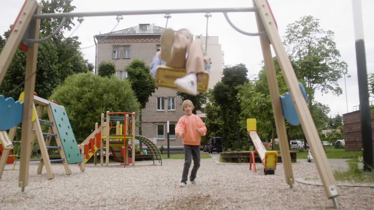 vista frontal de una niña con síndrome de down con capucha balanceándose en un columpio en el parque en un día ventoso. su amigo masculino empujándola mientras su amiga femenina toma una foto