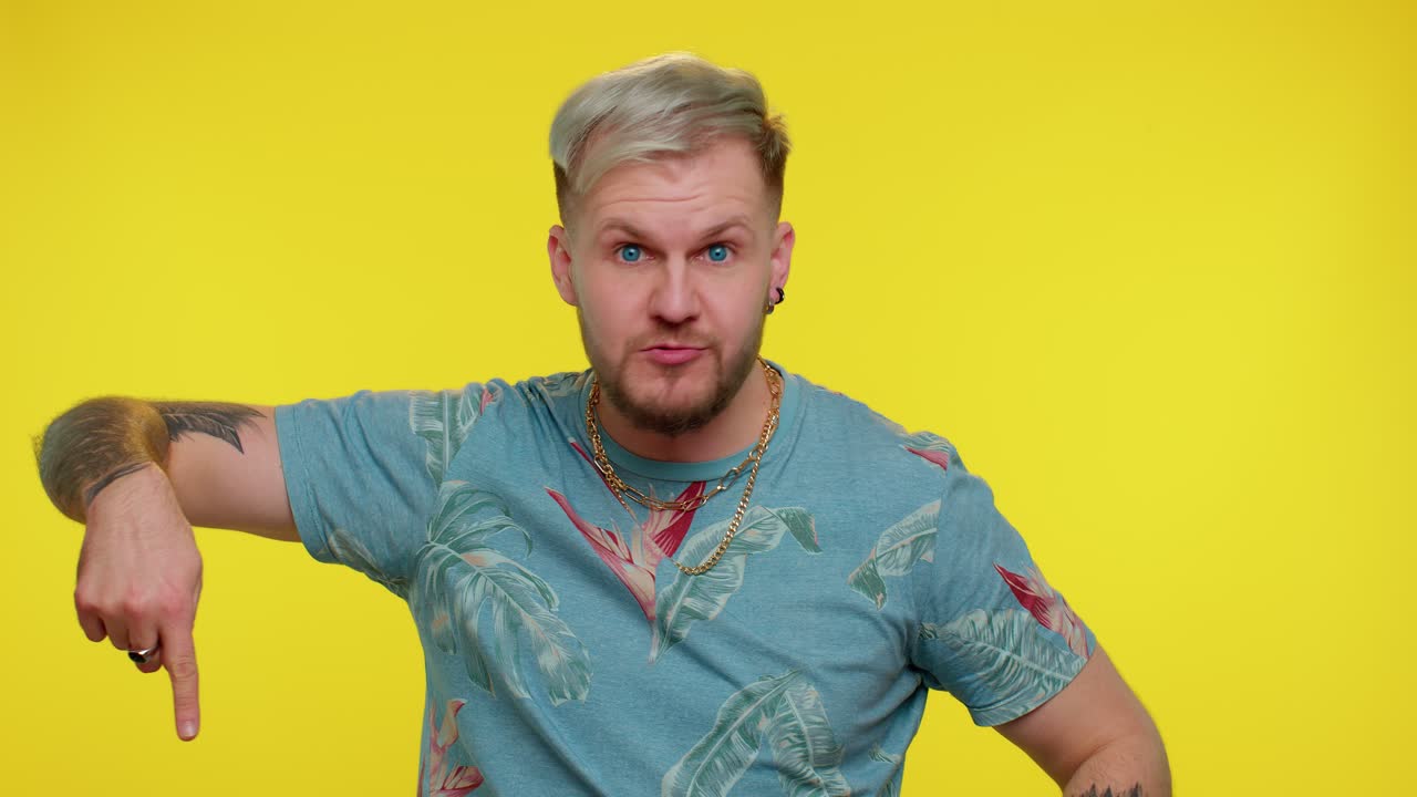 금발 머리카락을 가진 젊은 남자가 미소 짓고 엄지손가락을 올리고 열대 셔츠와 금 사슬을 입고 있습니다.