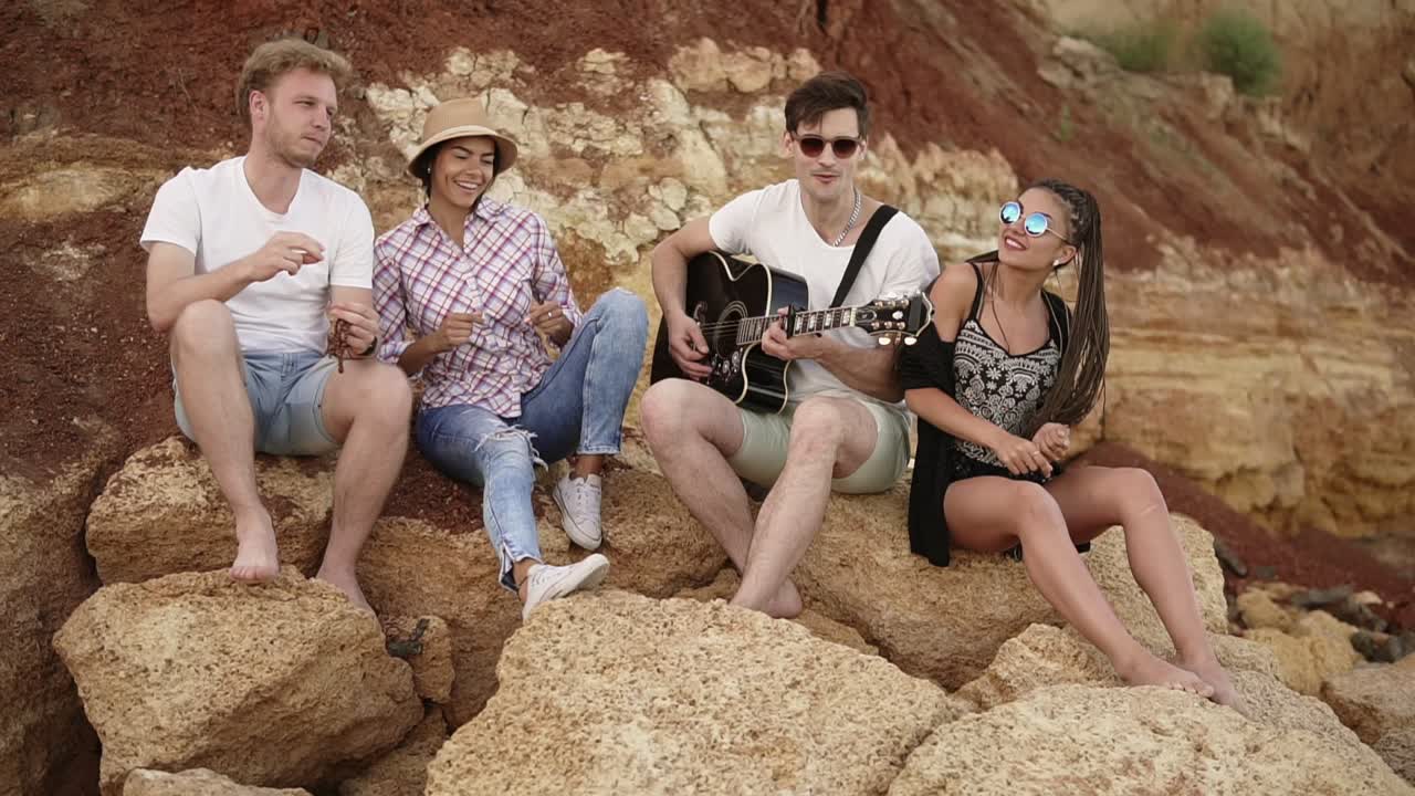 grupo de jóvenes amigos hipsters sentados en las rocas junto a la orilla del mar y tocando la guitarra, cantando canciones y bailando. cámara lenta