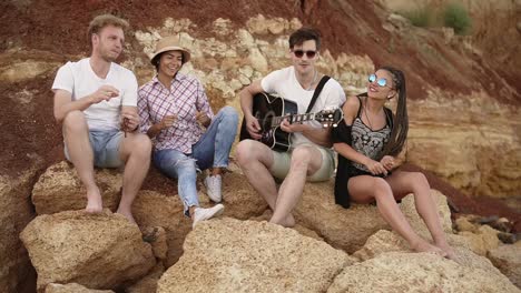 Group-of-young-hipster-friends-sitting-on-the-rocks-by-the-seashore-and-playing-guitar,-singing-songs-and-dancing.-Slow-Motion