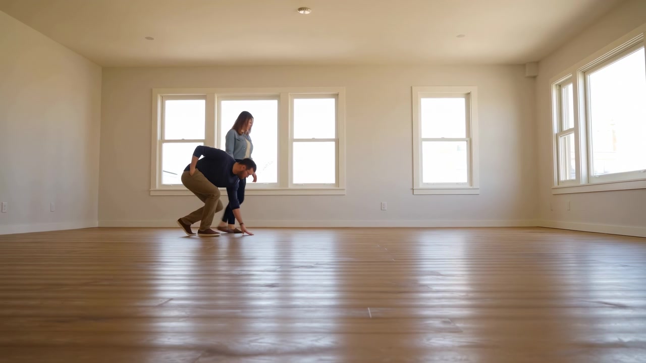 Man and woman move through large unfurnished interior, looking down as he reaches toward polished wooden boards before standing again, with large windows and pale walls in view.